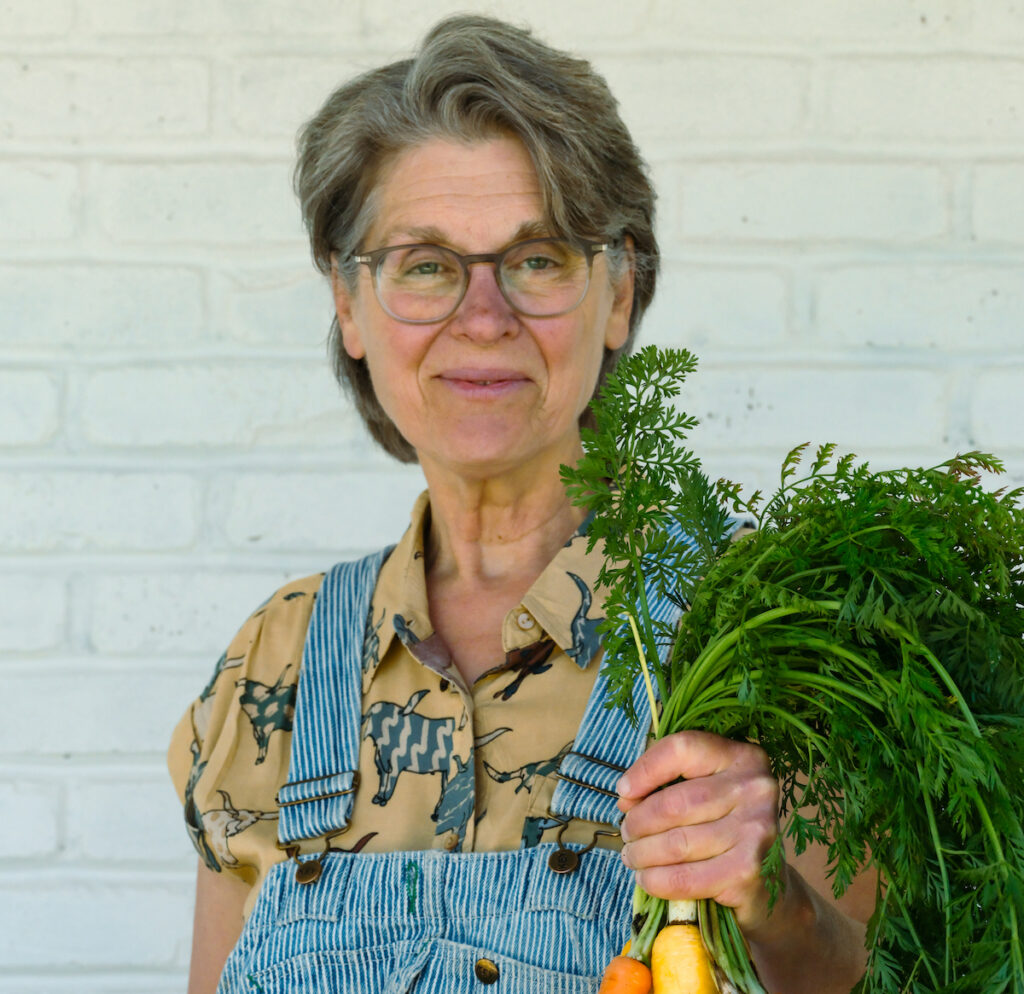 Woman in overalls holding a bunch of multicolored carrots.